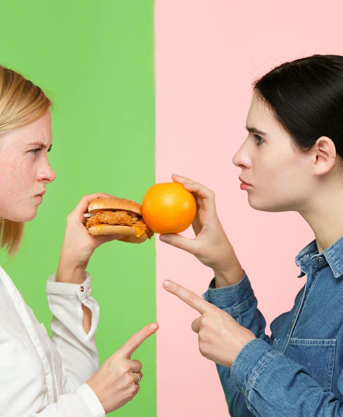 Diet. Dieting concept. Healthy useful food. Beautiful young women choosing between fruits and unhelathy fast food at studio. Human emotions and comparison concepts